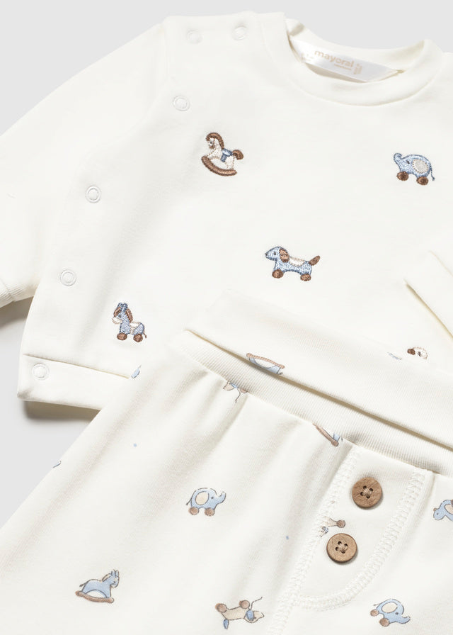 An off-white baby romper and matching pants set with small embroidered toy animal motifs, shown in a close-up flat-lay view with snap buttons and a buttoned pocket.