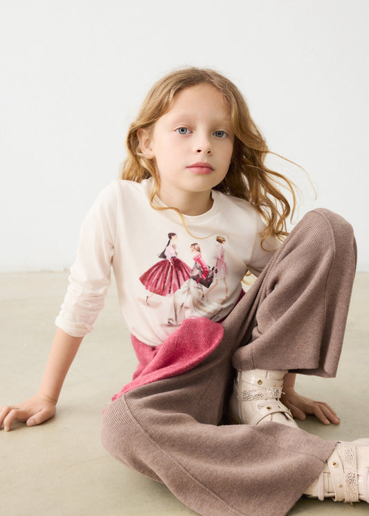 Young girl sitting on a light-colored floor wearing a white shirt with a graphic design and brown pants.