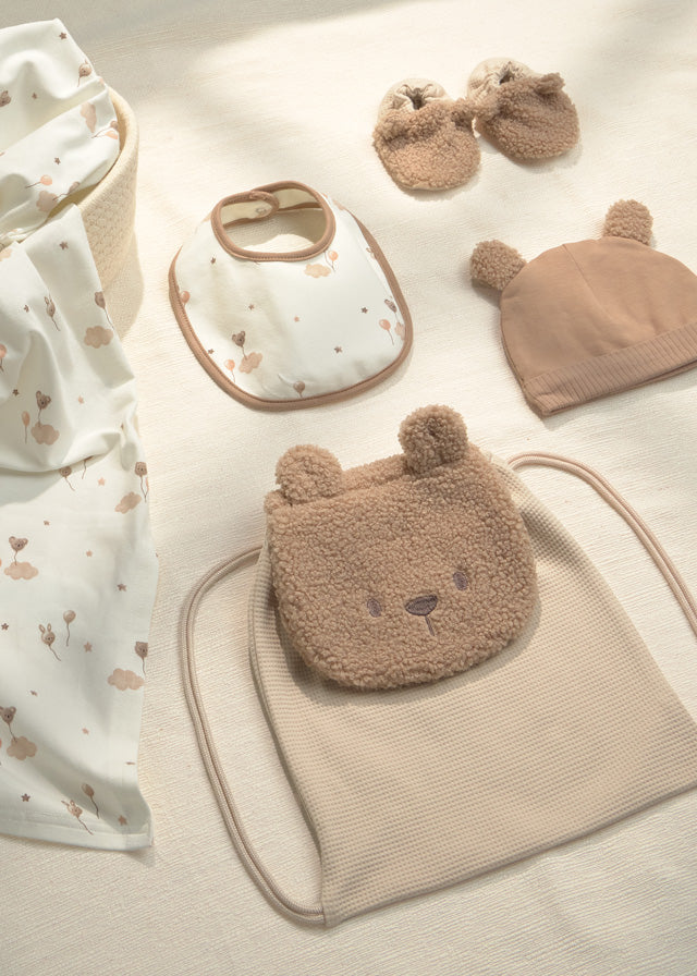 A coordinated baby gift set arranged on a light fabric surface, including a teddy-bear-face pouch, a ribbed drawstring bag, a bib, a knit hat, and a pair of bootie-style mittens. The palette is soft neutral tones with cream, beige, and tan.