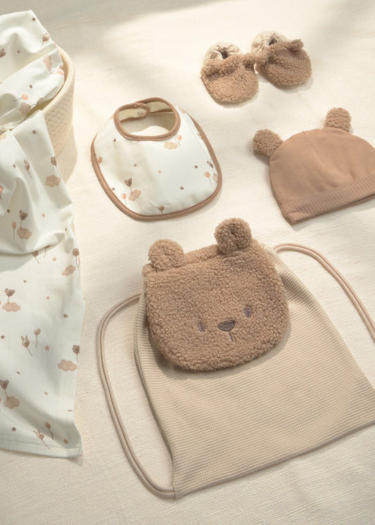 A coordinated baby gift set arranged on a light fabric surface, including a teddy-bear-face pouch, a ribbed drawstring bag, a bib, a knit hat, and a pair of bootie-style mittens. The palette is soft neutral tones with cream, beige, and tan.
