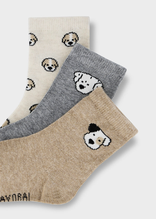 Three pairs of novelty socks with dog face patterns, displayed diagonally on a light gray background. The socks are in cream, heather gray, and heather beige, with the brand name 'MAYORAL' partially visible on the toe of the beige pair.