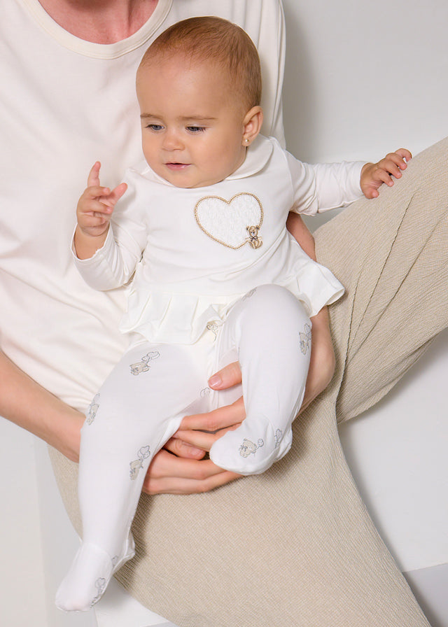 Baby in a white outfit with a heart design held by an adult against a neutral background