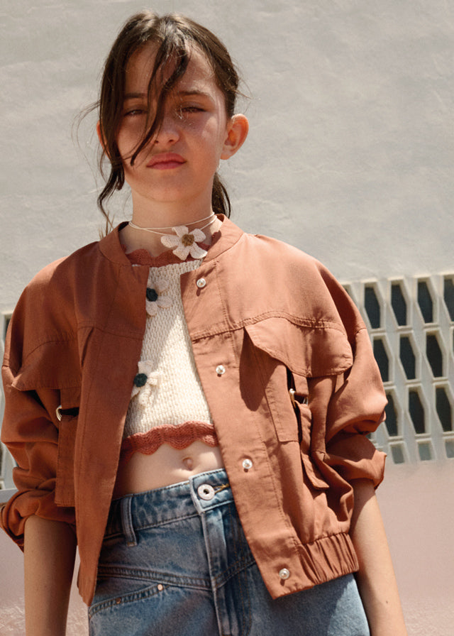 Person wearing a brown jacket over a white top with blue jeans against a textured wall.