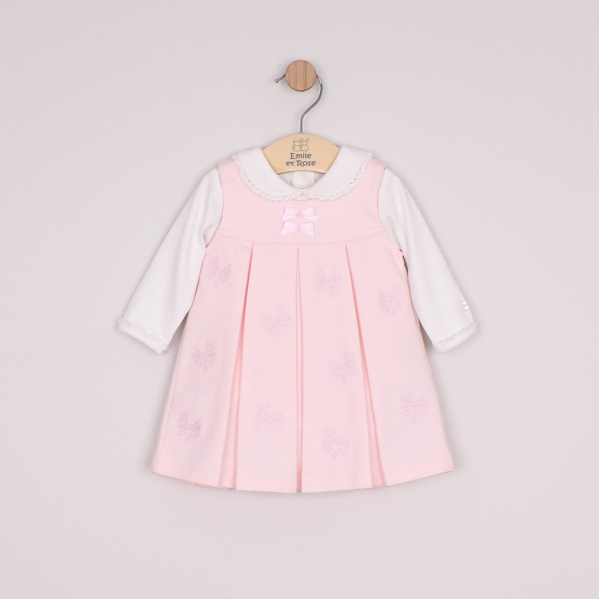 A baby girl's long-sleeve dress in light pink and white, featuring a pleated pink bodice with embroidered bow details, a white lace-trimmed collar, and a pink bow at the neckline.