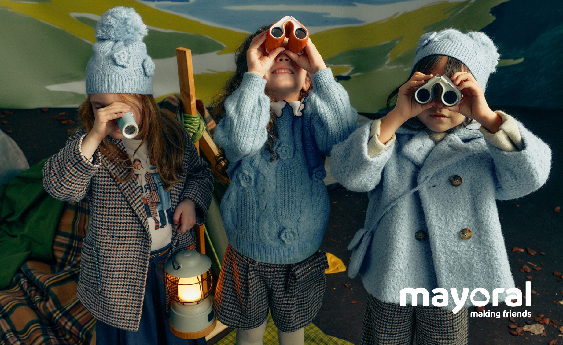 Three children dressed in cozy knit winter clothing pose outdoors, with two holding binoculars and one holding a lantern. The scene includes a visible 'mayoral making friends' logo in the lower right corner.