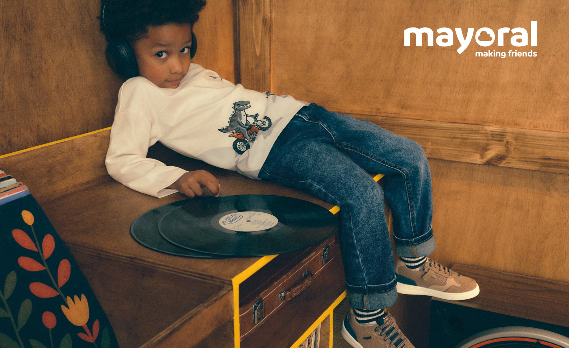A young child wearing a white long-sleeve shirt with a motorcycle graphic, blue jeans, and beige sneakers, sitting on a wooden bench next to a vinyl record. The Mayoral brand logo is visible in the top right corner.