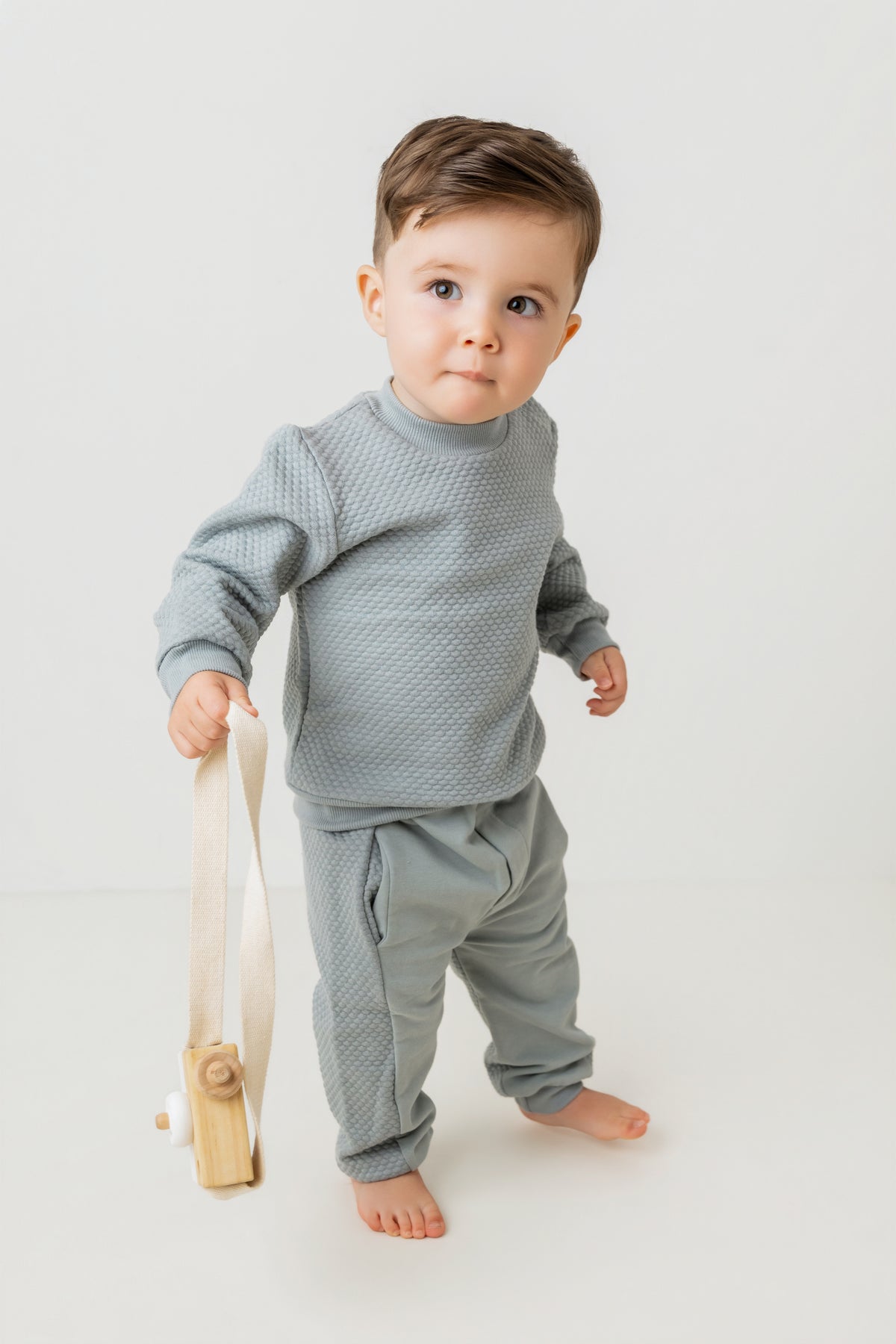 A toddler standing barefoot on a light gray studio floor, wearing a matching textured light gray long-sleeve top and jogger pants, while holding a small wooden toy with a cream strap.